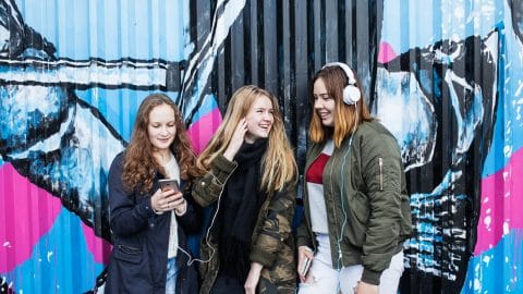 Kolme nuorta tyttöä nojailee iloisina graffitiseinään ja kuuntelee musiikkia kuulokkeilla puhelimistaan.
