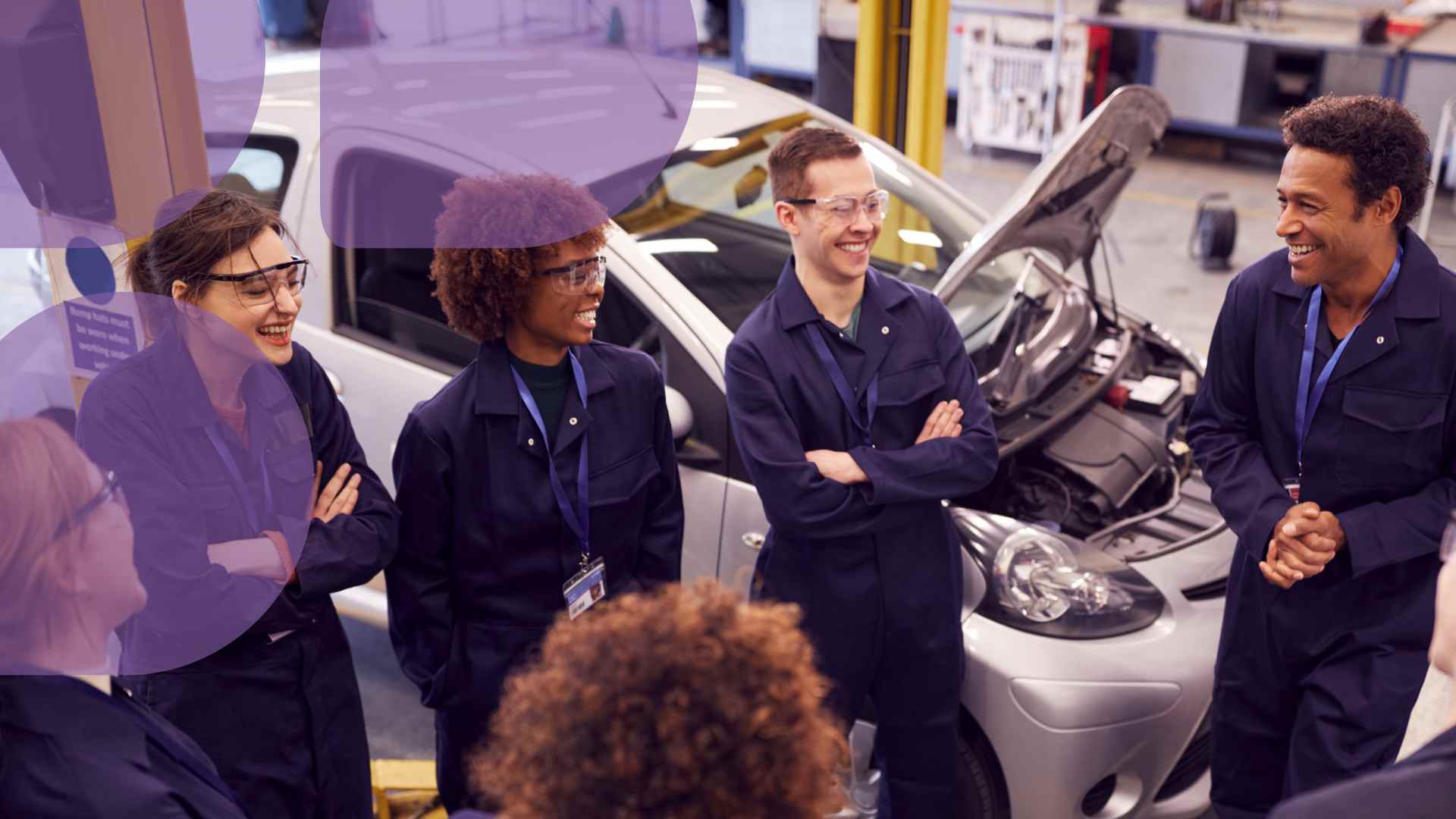 Ryhmä autoalan opiskelijoita seisoo tummansiniset työhaalarit päällä ringissä. Taustalla auto konepelti auki.